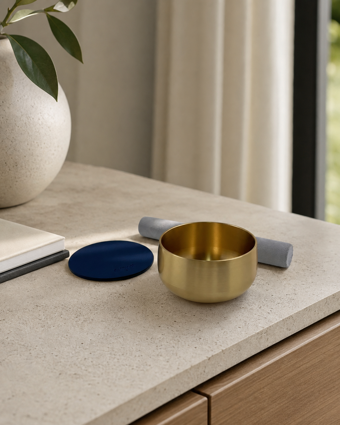 Brass singing bowl, navy pad and mallet on a stone counter by a window